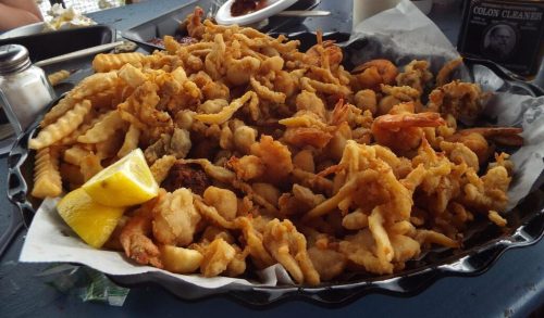 A large platter of fried seafood and fries, with a lemon wedge, on a table with condiments and a child in the background.