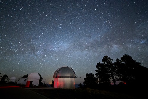 A clear night sky filled with stars and the Milky Way, with observatory domes illuminated by red light.