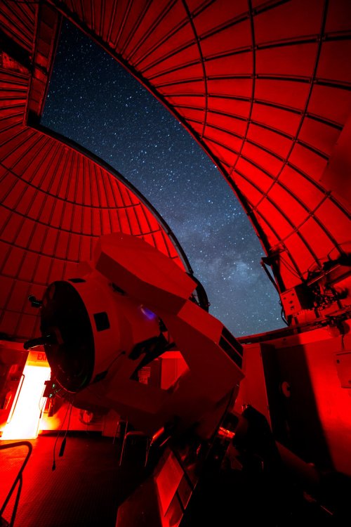 A telescope inside a red-lit observatory dome, with a starry night sky visible through the opening.