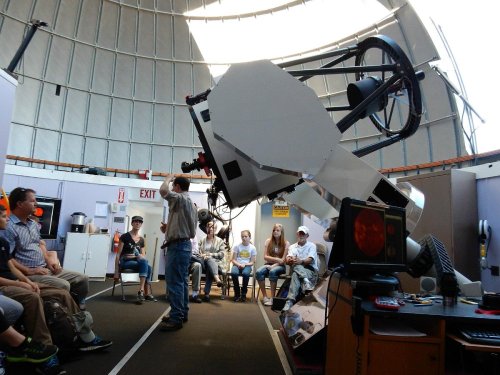 A group of people sits in an observatory, watching a presenter next to a large telescope.