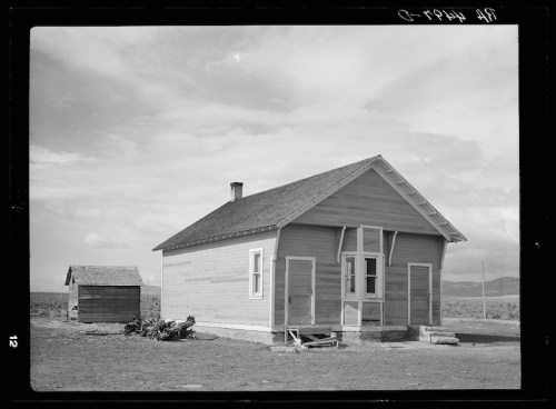 A simple wooden house with a sloped roof, set in an open landscape, with a smaller shed nearby under a cloudy sky.