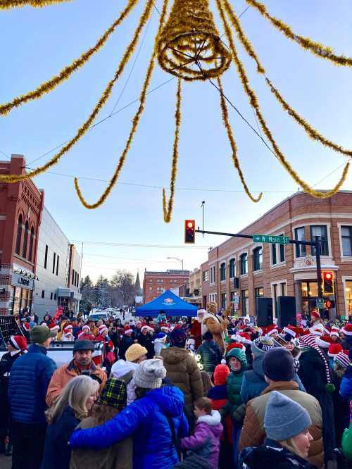 A festive crowd in winter attire gathers on a street decorated with holiday lights and a large golden ornament overhead.