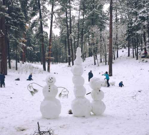 Three snowmen stand in a snowy forest, with people playing in the background among tall trees.