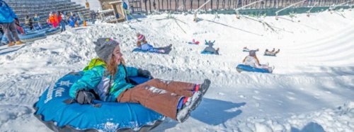 A child in a blue snow tube slides down a snowy hill, with others tubing in the background. Bright winter scene.