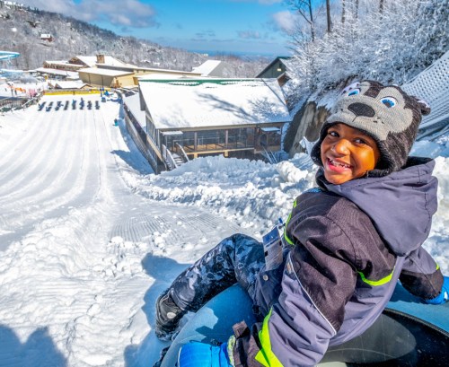 A smiling child in a winter hat sits on a snow tube, ready to slide down a snowy hill at a winter park.