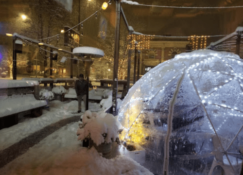 A snowy scene at night featuring a lit igloo structure and a person standing nearby, surrounded by winter decor.