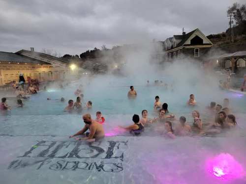 A crowded hot springs pool with steam rising, colorful lights, and people enjoying the warm water on a cloudy day.