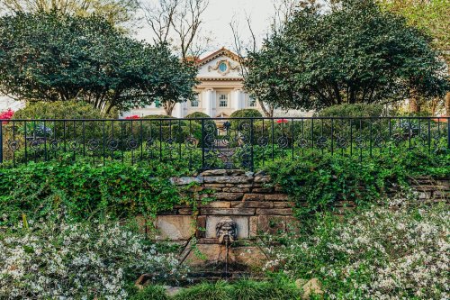 A lush garden with a stone fountain, surrounded by greenery and flowers, in front of a grand building.