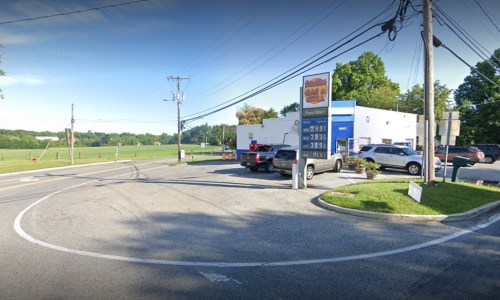 A street view of a gas station at a curve, with parked cars and green fields in the background.