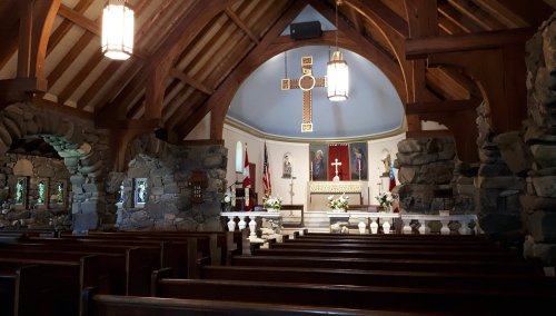 Interior of a church featuring wooden beams, stone walls, a large cross, and rows of wooden pews.