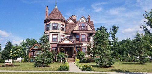 A large, ornate Victorian-style house with a turret, surrounded by lush greenery and colorful flowers.