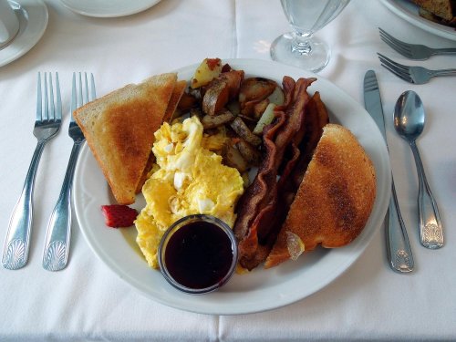 A plate of breakfast featuring scrambled eggs, bacon, toast, potatoes, and a side of jam, garnished with a strawberry.