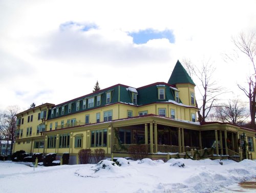 A large yellow Victorian-style building with a green roof and a turret, surrounded by snow and a cloudy sky.