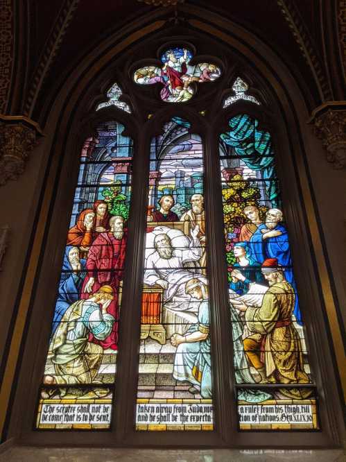 Stained glass window depicting a religious scene with figures gathered around a central figure, vibrant colors and intricate details.