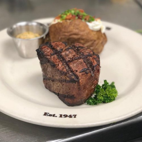 A grilled steak with a crosshatch pattern, served with a baked potato and a small cup of sauce on a plate.
