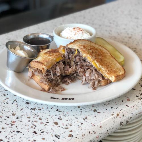 A plate with a toasted sandwich filled with pulled pork, served with a side of coleslaw, pickles, and dipping sauces.