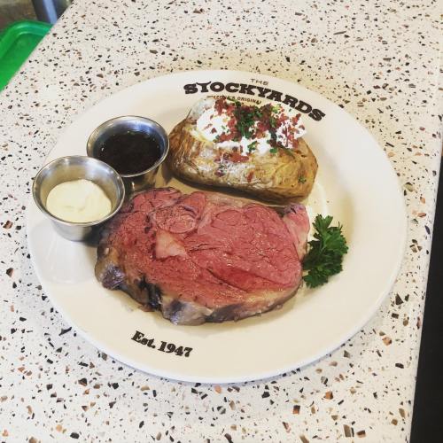 A plate featuring a juicy prime rib, baked potato with toppings, and dipping sauces, served at The Stockyards.