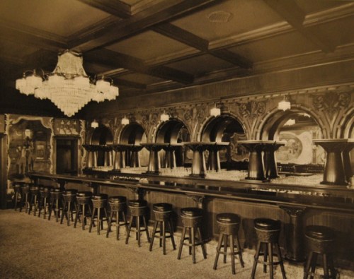 A vintage bar interior featuring a long counter, high stools, ornate arches, and a chandelier.