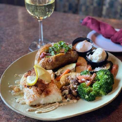 A plate of grilled fish with lemon, mixed vegetables, and a baked potato, accompanied by a glass of white wine.