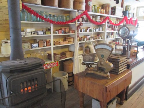 A vintage store interior featuring shelves of jars and cans, a scale, and a wood stove, decorated with red garlands.