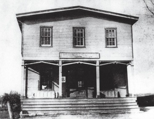Black and white photo of a two-story wooden building with a porch and multiple windows.