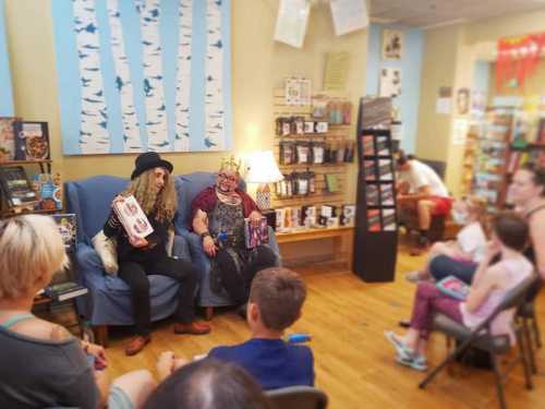 Two people in costumes hold books while seated on a couch, engaging with an audience in a cozy bookstore setting.