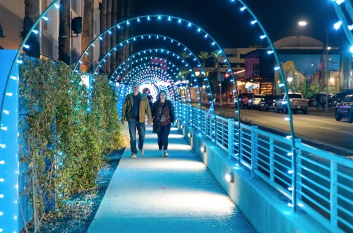 A couple walks through a brightly lit, blue-lit archway at night, surrounded by vibrant city lights.
