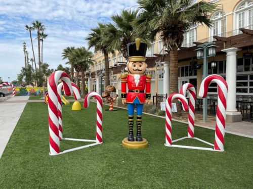 A festive scene featuring a nutcracker statue surrounded by large candy canes on a grassy area with palm trees.