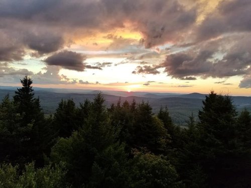 A scenic sunset over a mountainous landscape, framed by lush green trees and a colorful sky filled with clouds.