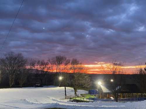 A winter landscape at sunset, with snow-covered ground and trees silhouetted against a colorful sky.