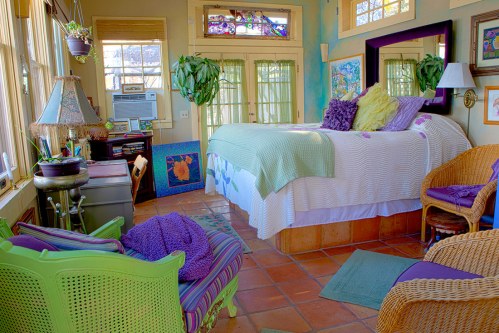 Cozy bedroom with a bed, colorful decor, plants, and natural light from large windows. Warm, inviting atmosphere.