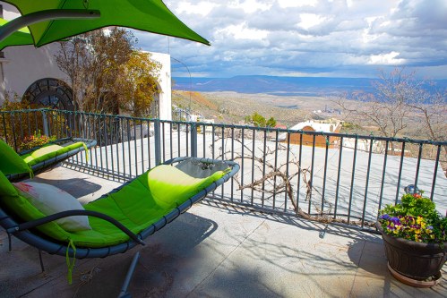 A balcony with green lounge chairs overlooks a scenic valley under a cloudy sky. Potted plants add color to the space.