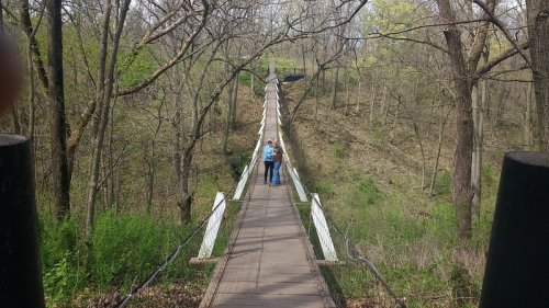 A couple walks hand-in-hand across a suspension bridge surrounded by trees and greenery.