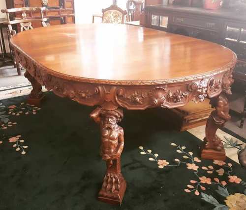 A large, ornate wooden dining table with intricately carved legs featuring figures, set on a floral-patterned rug.