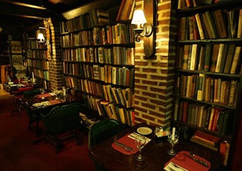 Cozy restaurant interior with wooden tables, green chairs, and a wall of bookshelves filled with old books.