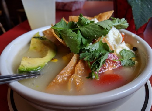 A bowl of soup topped with avocado, tortilla strips, cilantro, and diced tomatoes, served on a red table.
