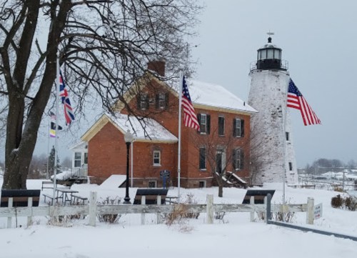 A snowy scene featuring a brick building and a lighthouse, both adorned with American flags, surrounded by winter landscape.
