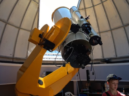 A large telescope inside an observatory dome, with a young person standing nearby, observing the equipment.