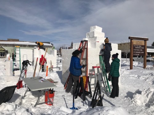 People working on snow sculptures using ladders and tools in a snowy outdoor setting, with a sign in the background.