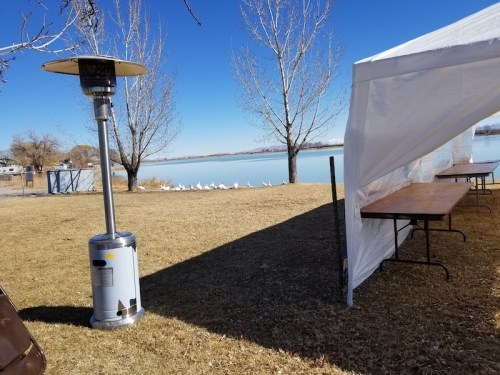 A lakeside scene with a patio heater, empty tables under a tent, and trees beside calm water.