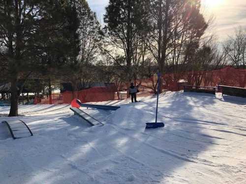 A snowy landscape with ski ramps and a person standing near a flag, surrounded by trees and a safety fence.