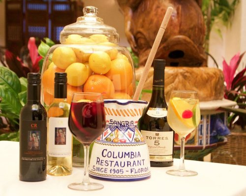 A table display featuring wine bottles, a pitcher of sangria, and a jar of oranges, surrounded by tropical plants.