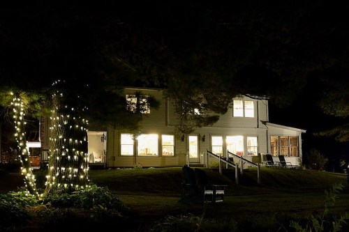 A cozy house at night, illuminated by warm lights, surrounded by trees and a well-lit porch.