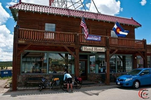 A rustic wooden hotel with flags, featuring a cyclist by parked bikes in front of the Hand Hotel in Colorado.