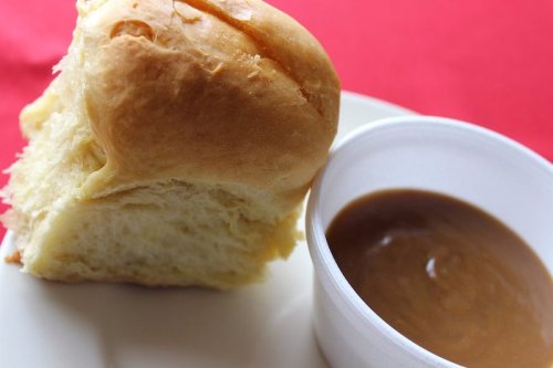 A soft bread roll next to a small cup of brown sauce on a white plate, set against a red background.
