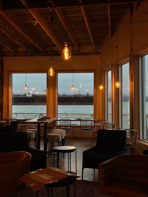 Cozy interior with hanging lights, tables, and a view of the water and distant ships through large windows.
