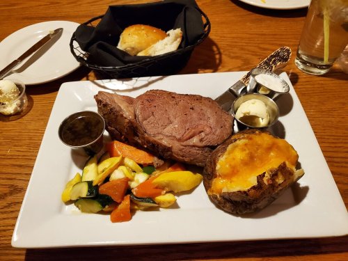 A plate with prime rib, baked potato with cheese, mixed vegetables, and a roll, served on a wooden table.