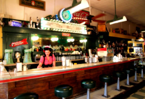 A vintage diner with a woman in a pink shirt serving at the counter, featuring retro decor and green stools.