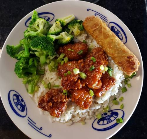 A plate of rice topped with sweet and sour chicken, broccoli, and an egg roll on the side.