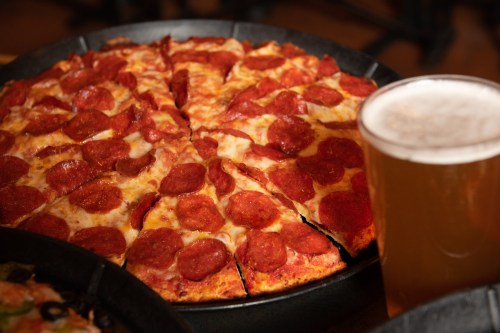 A freshly baked pepperoni pizza sliced into pieces, next to a glass of beer on a wooden table.